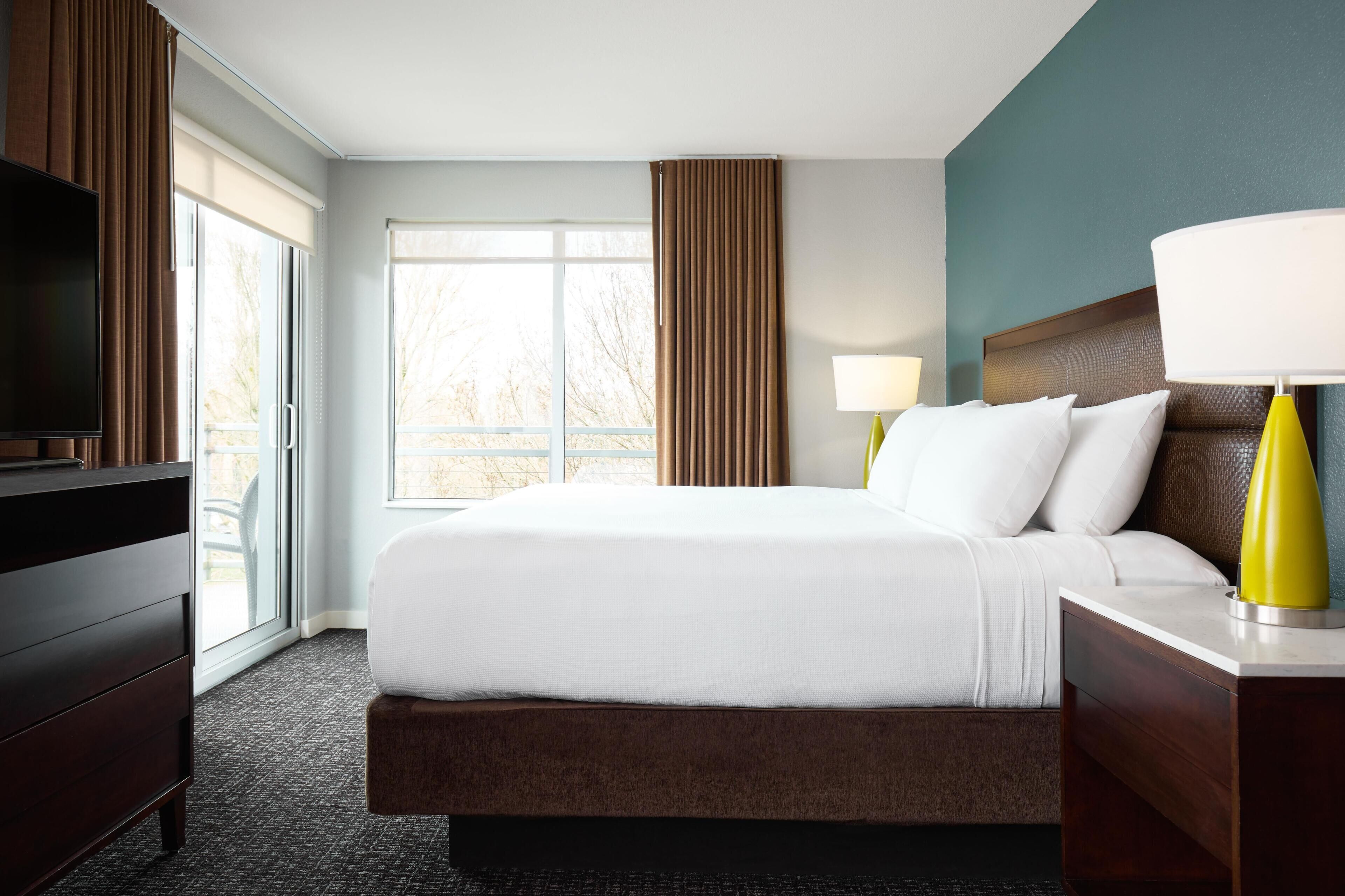 A hotel room with a large bed, a TV, a desk with a lamp, and a window with curtains.