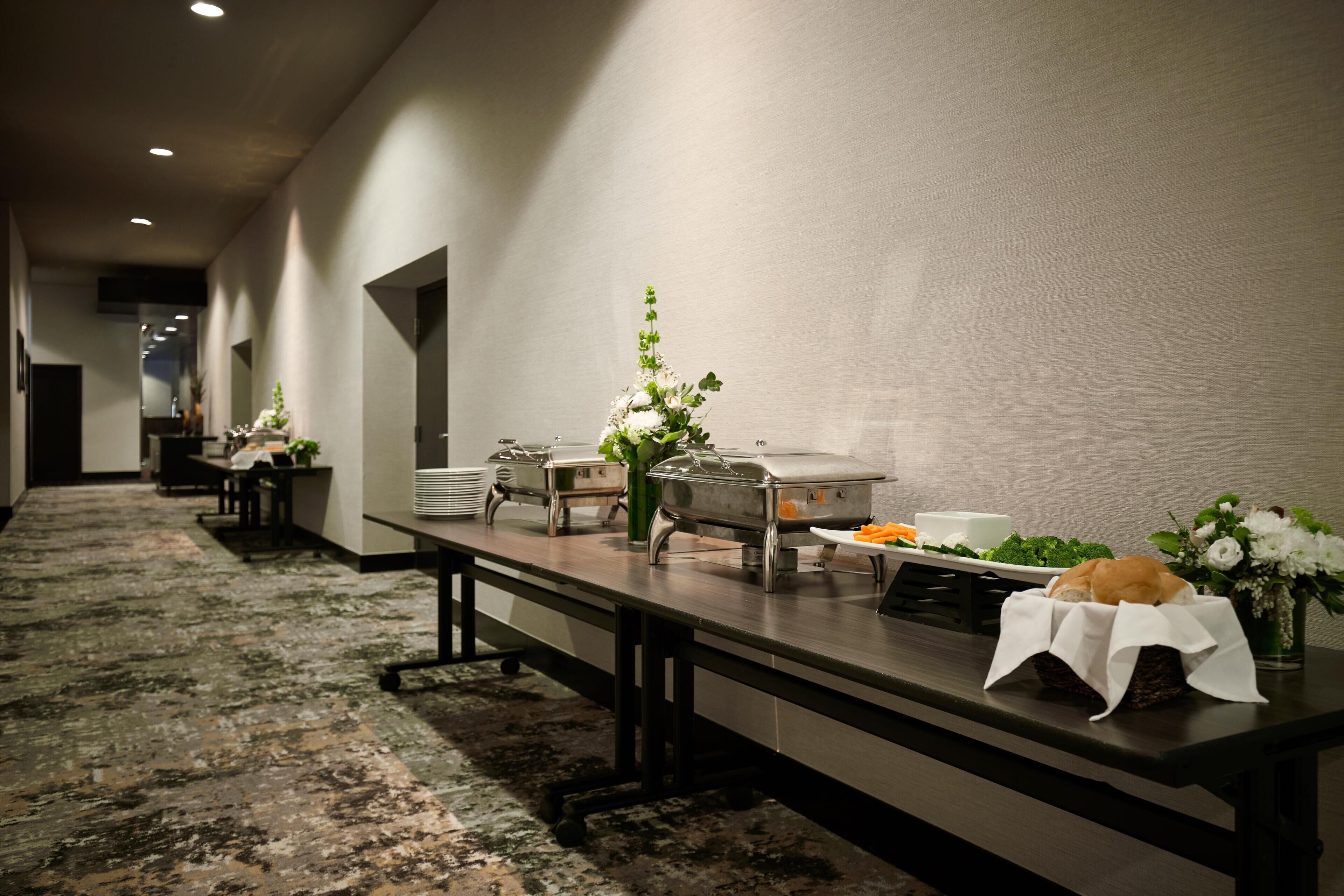 Una larga mesa de banquete con equipo de servicio de alimentos, un jarrón con flores y una cesta con pan.