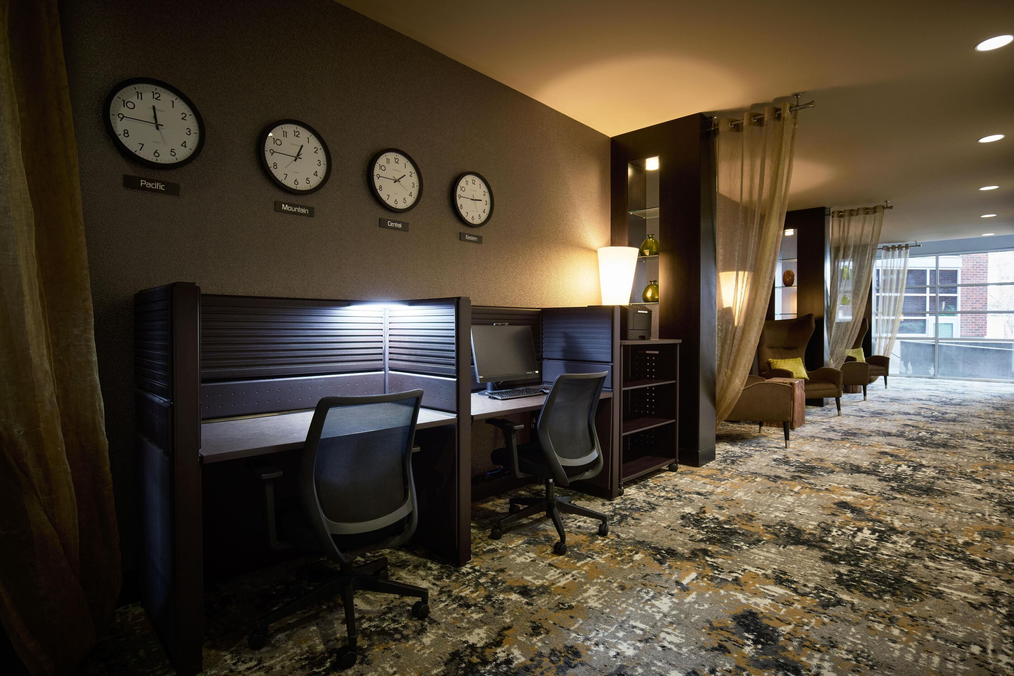 A hotel room with a work area featuring two desks and chairs, a large window with curtains, and three clocks on the wall indicating Pacific, Mountain, and Central time zones.