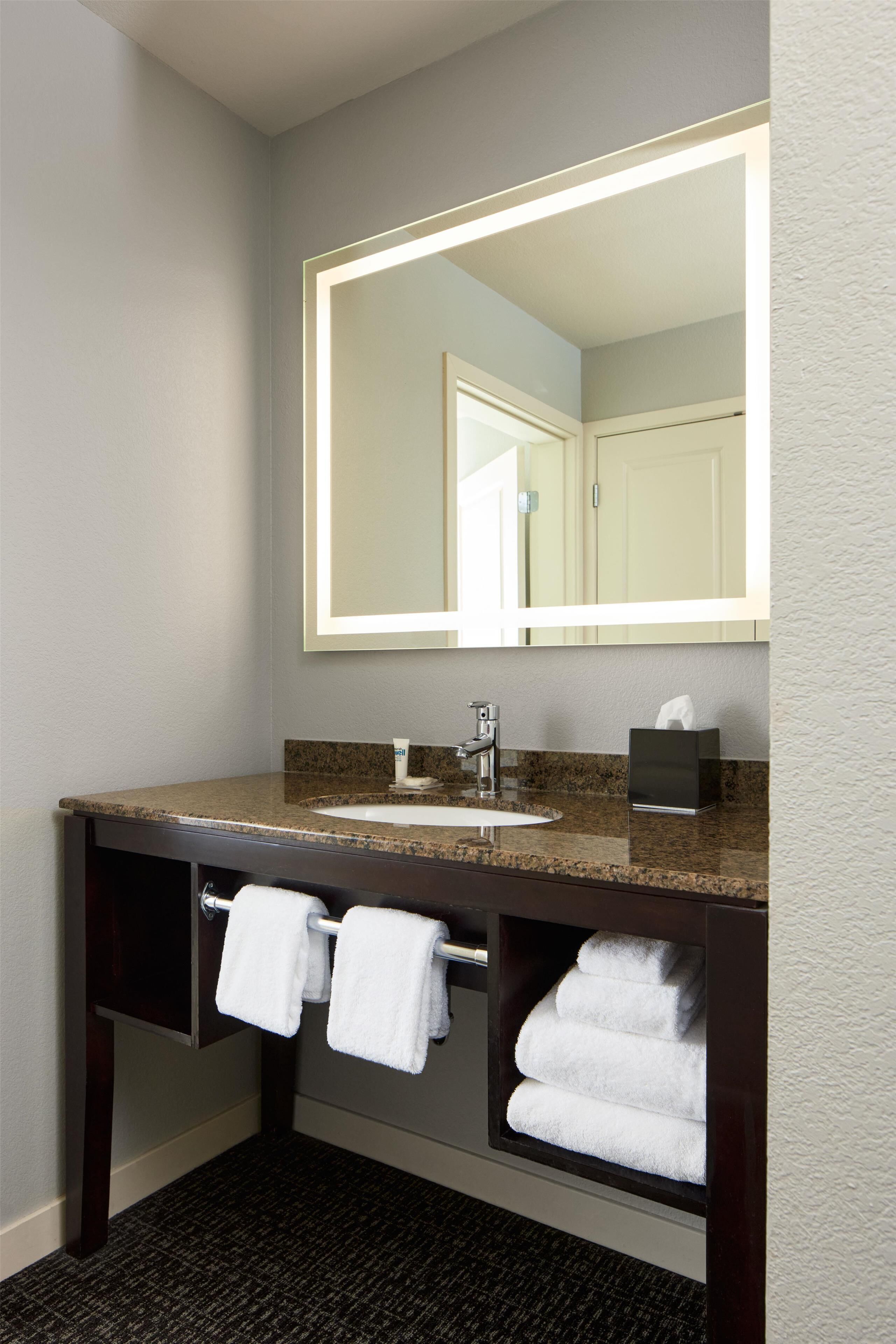 A bathroom with a granite countertop sink, a mirror with integrated lighting, a towel rack, and a built-in towel shelf.