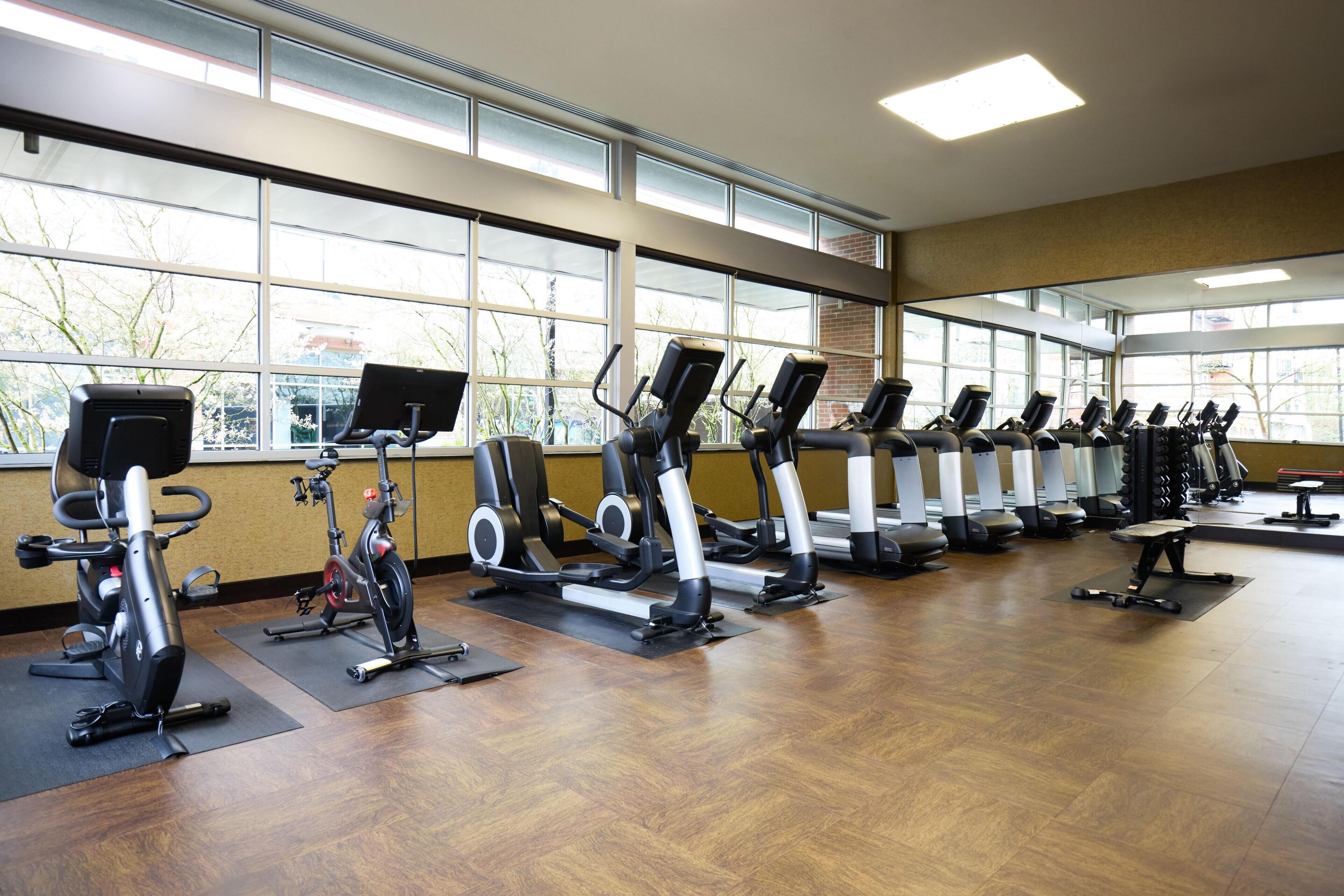 A gym with multiple cardio machines, large windows, and a wooden floor.