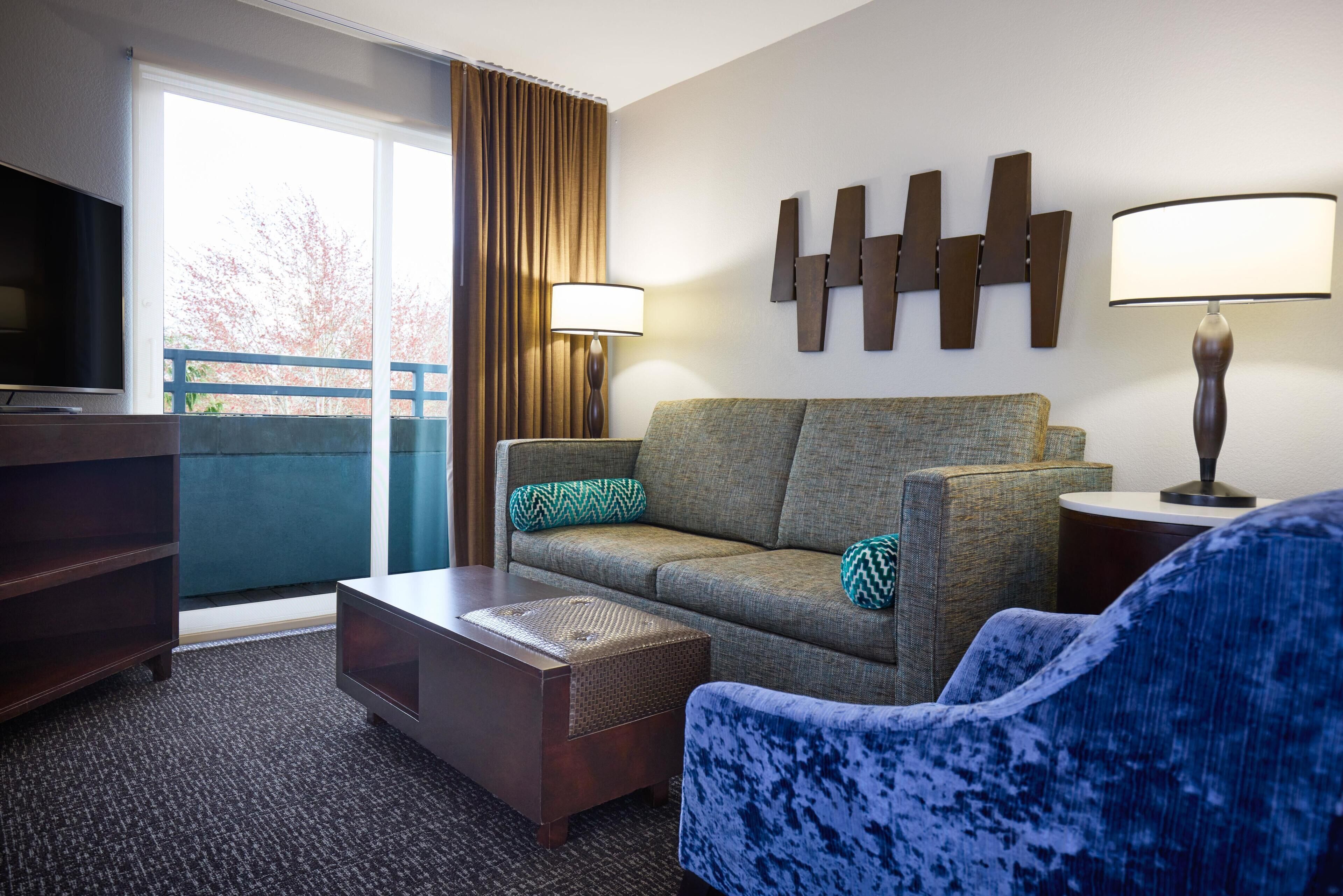 Une chambre d’hôtel équipée d’un canapé, d’un fauteuil, d’une télévision, d’une lampe et offrant une vue sur l’extérieur.