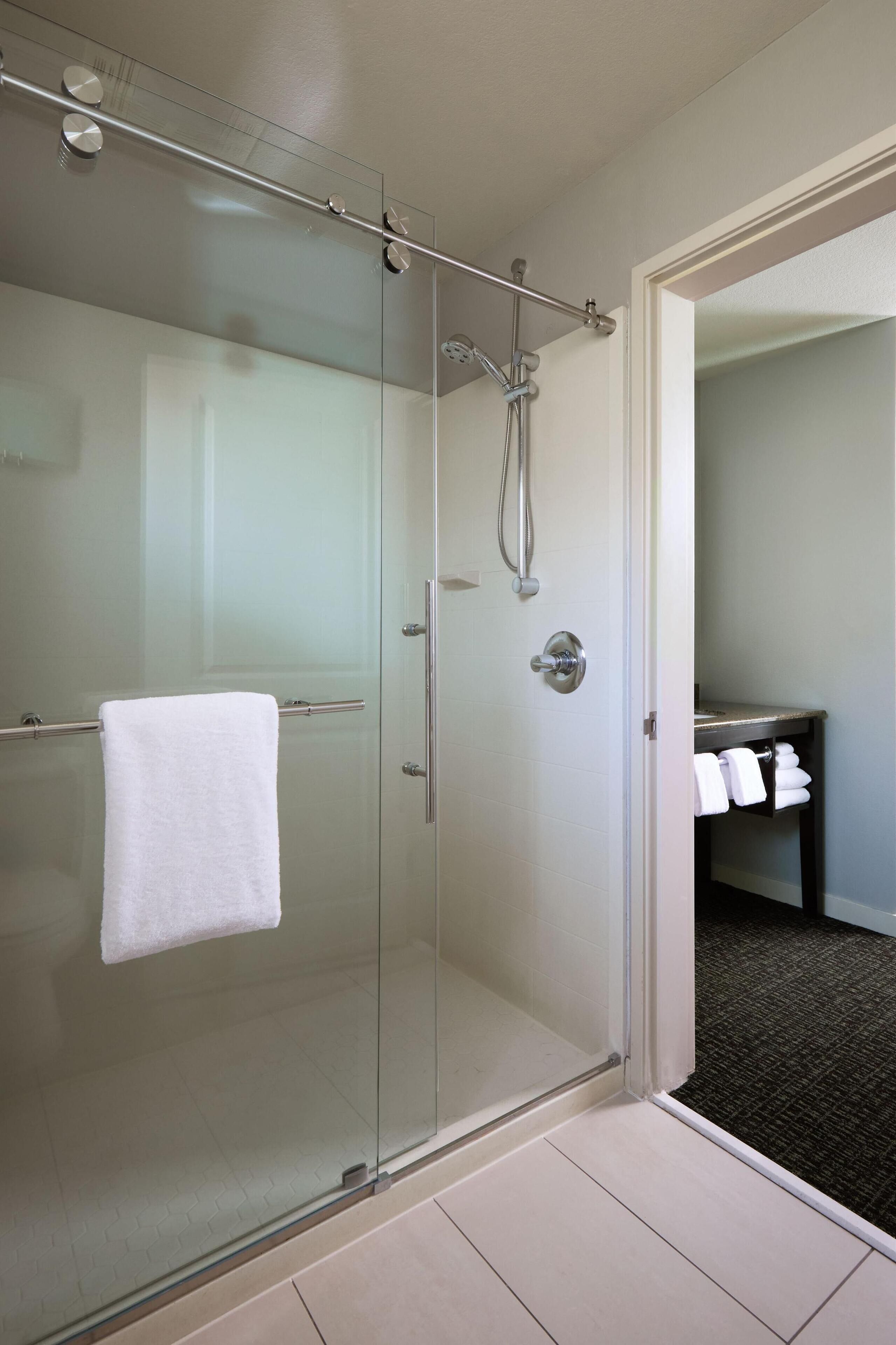 Un baño moderno con cabina de ducha de vidrio, una toalla blanca colgada en la puerta y un lavabo con toallas a la vista.