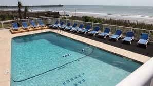 A pool with clear water, surrounded by blue lounge chairs, and a view of the beach and ocean.