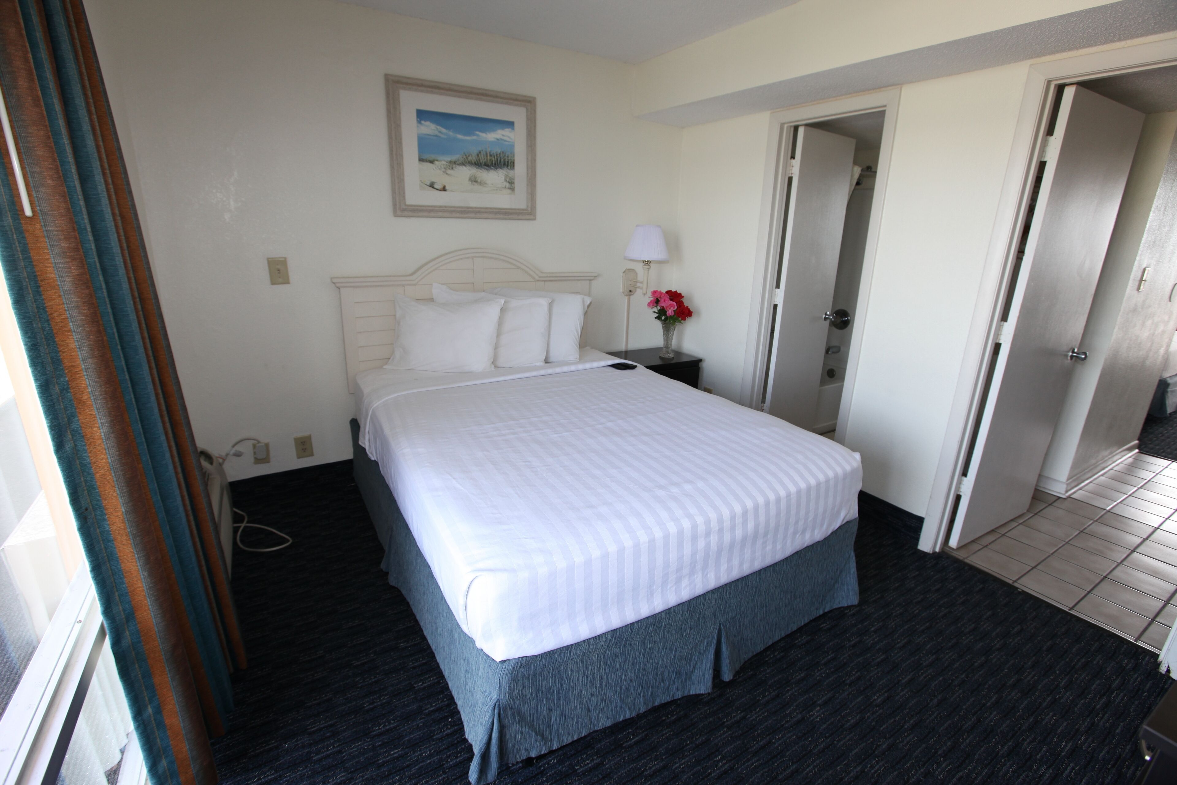 A hotel room with a large bed, a nightstand with a lamp, a framed picture on the wall, and a door leading to another room.