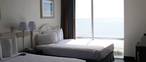 Habitación de hotel con dos camas, un escritorio, una silla, una lámpara y un amplio ventanal con vista al mar.
