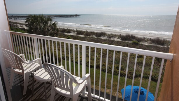 A balcony with white chairs and a table, overlooking a beach with a pier, sandy dunes, and a clear sky.