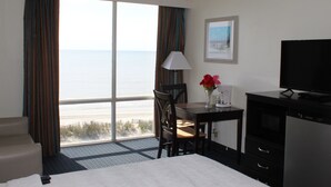 A hotel room with a large window, a TV, a desk with a lamp and flowers, and a bed.
