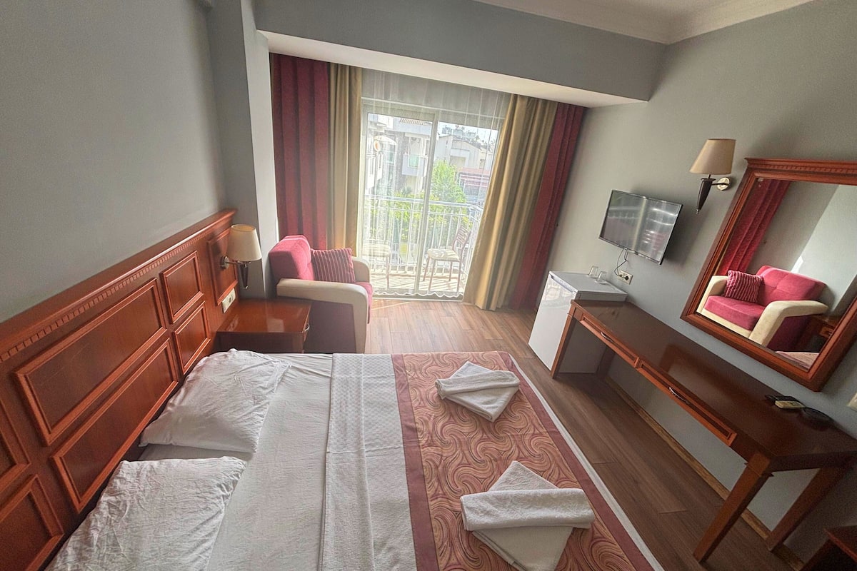 Une chambre d’hôtel équipée d’un lit, d’un bureau, d’une chaise, d’une télévision et d’un balcon avec des rideaux.