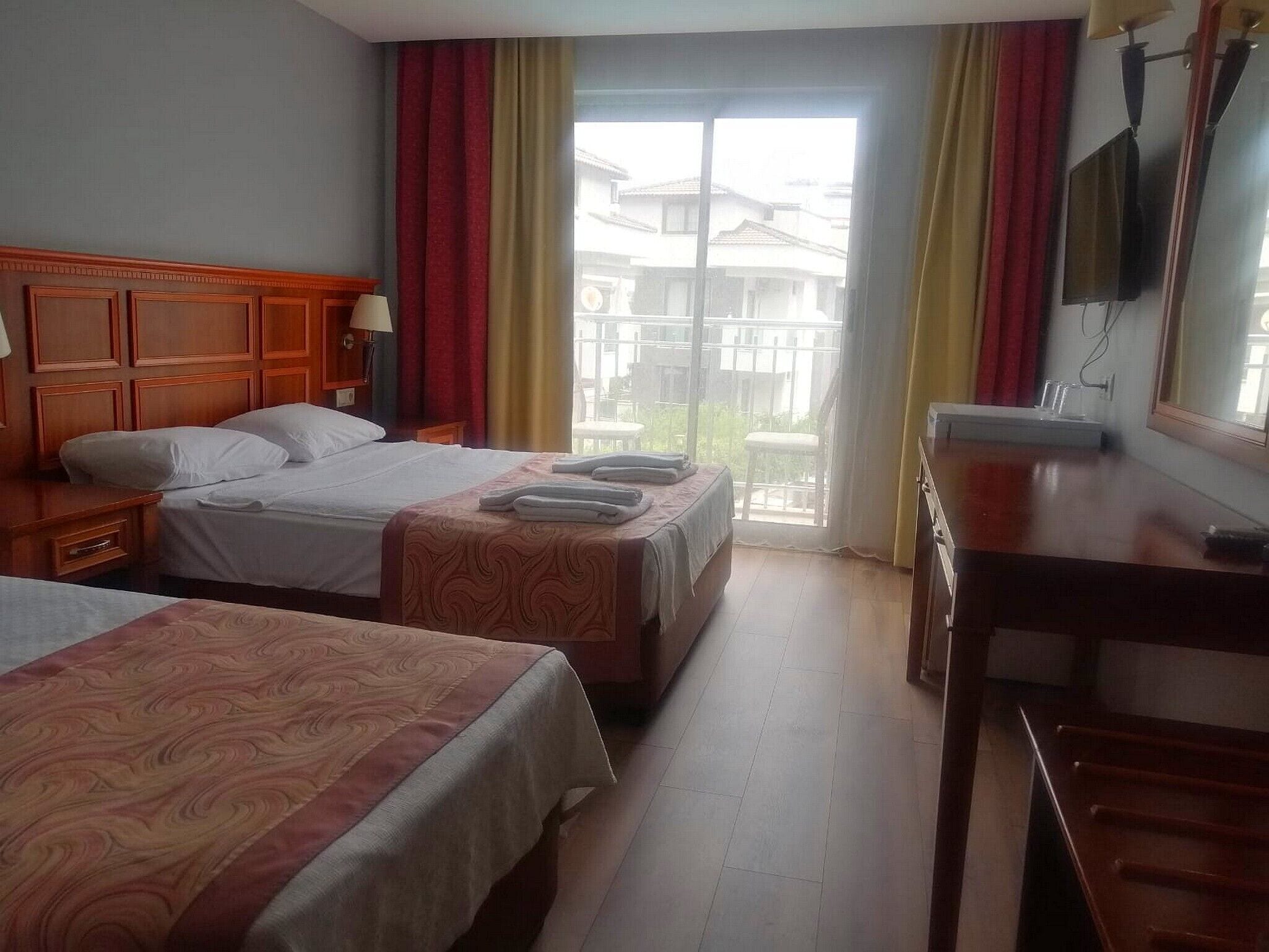 A hotel room with two beds, a wooden headboard, a desk, a TV, and a balcony with a view of buildings.