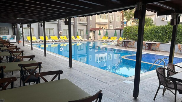 Uma área junto à piscina com mesas e cadeiras, uma piscina e um edifício ao fundo.