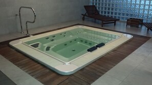 Un espace spa doté d’un grand bain à remous extérieur, de chaises longues en bois et offrant une vue à travers une grande fenêtre.