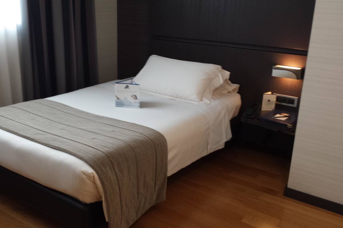 Een netjes opgemaakt bed met een witte deken en een grijze plaid, een nachtkastje met een lamp, en een houten vloer.