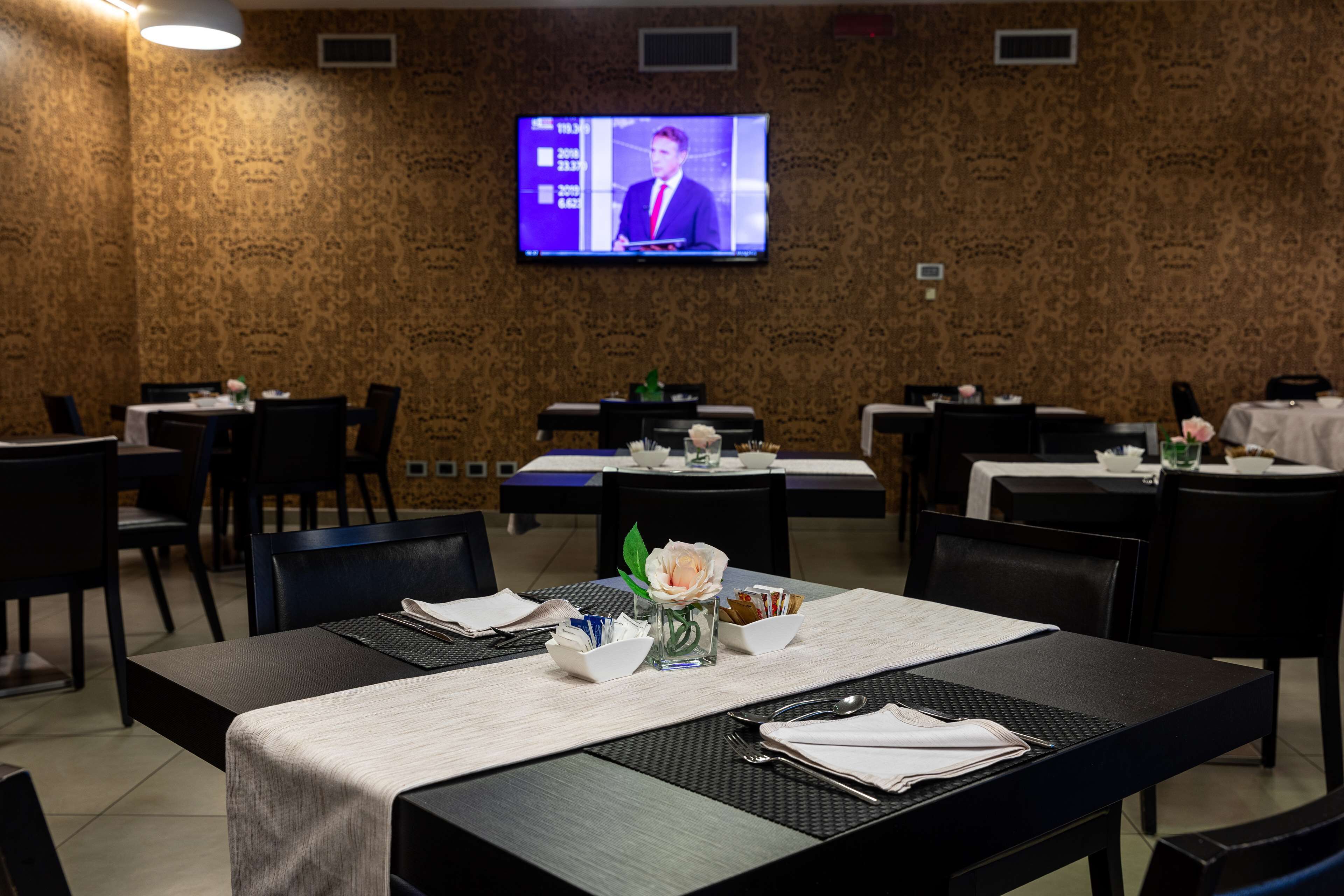 A conference room with a television displaying a news broadcast, tables with white tablecloths, and black chairs.