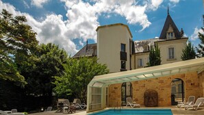 Una piscina con sillas de descanso, un área de estar techada y un edificio con una torre al fondo.