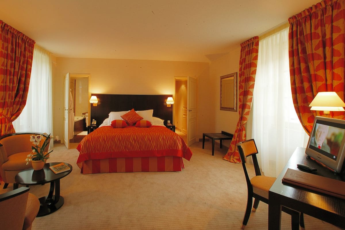 A hotel room with a large bed, a desk with a computer, a chair, a small table with a vase of flowers, and a lamp.