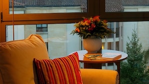 Una habitación con un ventanal grande, una silla con un cojín a rayas, una mesita con un jarrón de flores y vistas a edificios y colinas.