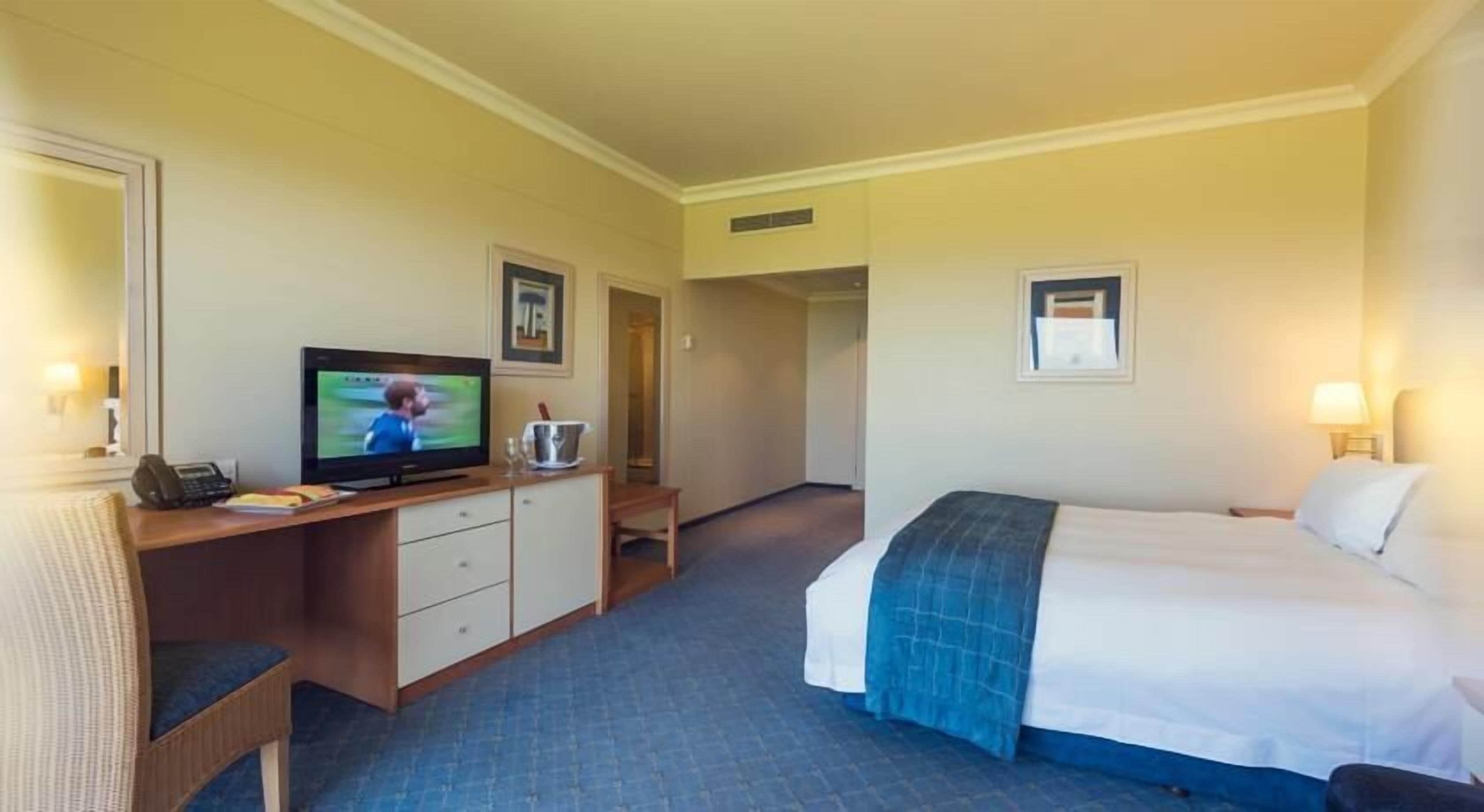 A hotel room with a bed, a desk with a television, a chair, a bedside table, and a lamp.