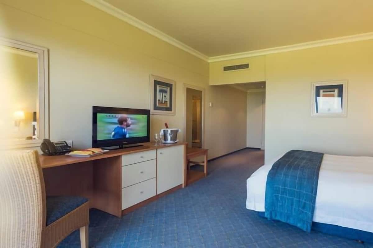 A hotel room with a bed, a desk with a television, a chair, a bedside table, and a lamp.