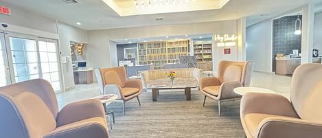 A modern lobby with a central seating area, a reception desk, and a kitchen in the background.