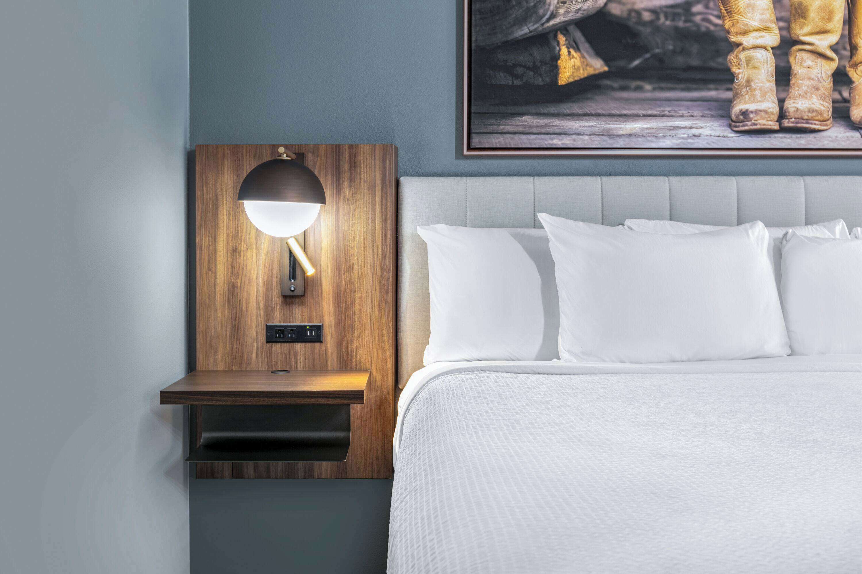 En pænt redt seng med hvide sengetøj, et trænatbord med en lampe, og et indrammet kunstværk over sengen.