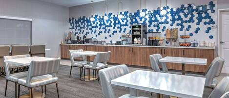 A modern dining area with white tables and chairs, a kitchen counter with various appliances, and a wall decorated with blue hexagonal patterns.