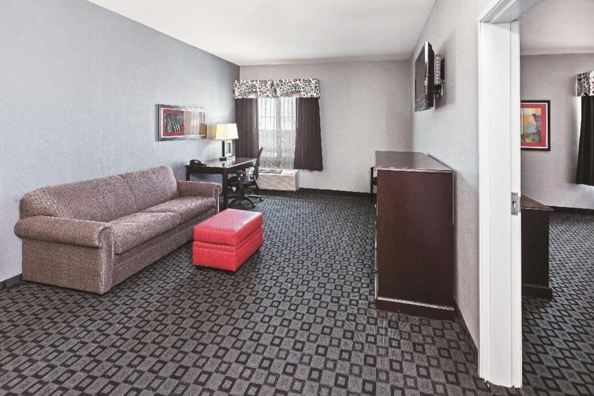 Une chambre d’hôtel comprenant un canapé, une petite table, un pouf rouge, un bureau, une chaise, un téléviseur et une moquette à motifs.