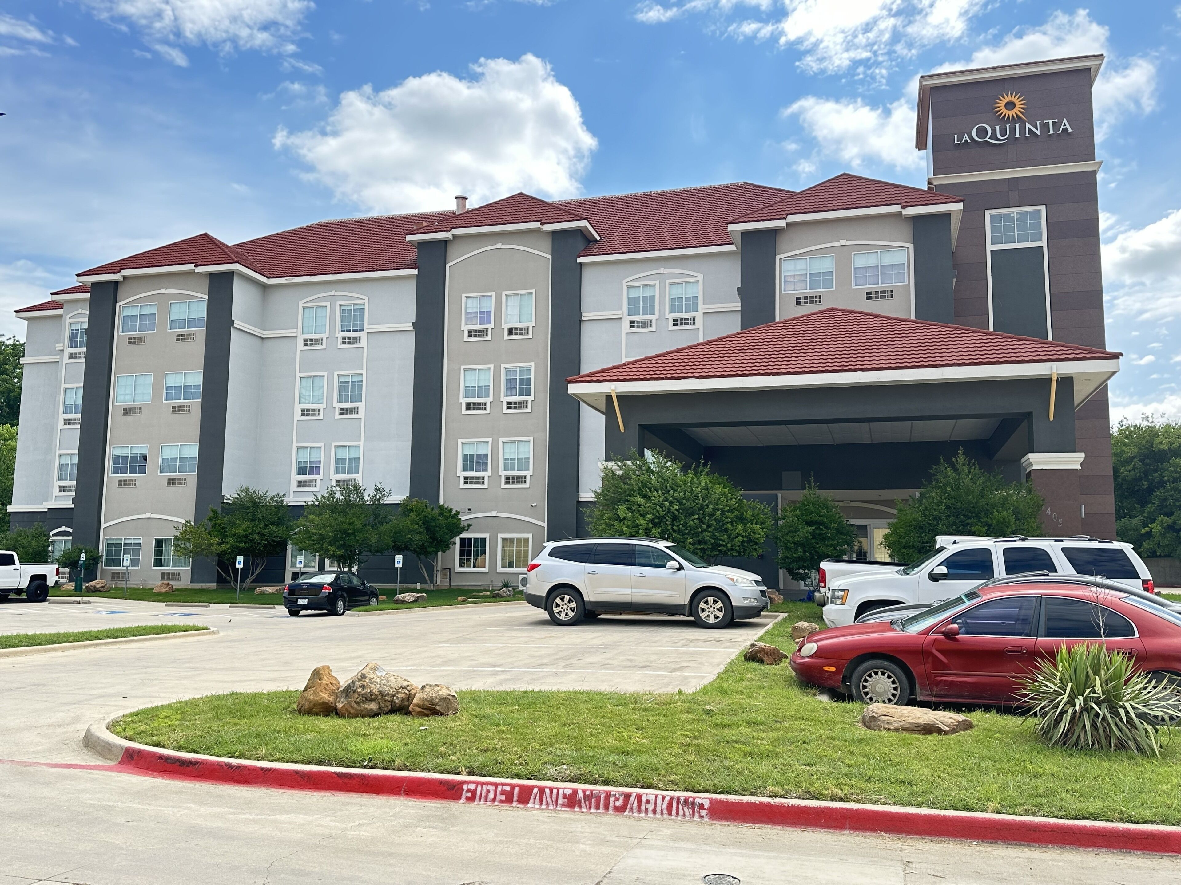 A multi-story hotel with a red-tiled roof, a covered entrance, and a parking lot with several cars.