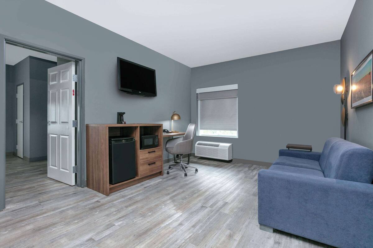 Un salon moderne avec un canapé bleu, un meuble en bois contenant un four à micro-ondes, un bureau avec une chaise, un téléviseur à écran plat fixé au mur et une fenêtre avec des stores.