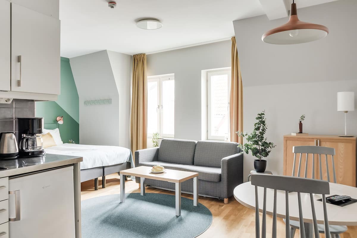 A modern hotel room with a bed, sofa, dining table, and kitchenette.