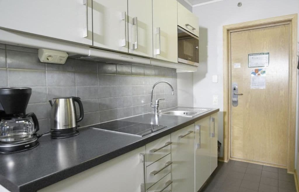 A kitchen with a sink, a coffee maker, and a kettle. There is a wooden door and a cabinet with a handle.