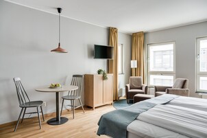 Et hotelværelse med en seng, to stole, et lille bord, et fjernsyn og et vindue med gardiner.