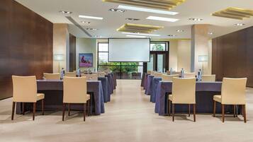 A conference room with rows of tables and chairs, a projector screen, and a large window with a view of greenery.