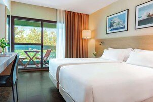 Habitación de hotel con dos camas, un escritorio, una silla, una ventana con cortinas y dos cuadros en la pared.