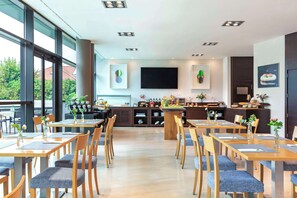 A modern dining area with wooden tables and chairs, a central bar area with various items, and a large window providing a view of the outdoors. - NH Gijón (Gijon)