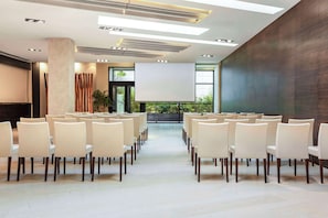 Un salón de conferencias con filas de sillas orientadas hacia una pantalla grande, iluminación moderna y ventanas amplias.