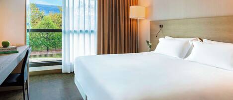 Habitación de hotel con una cama grande, un escritorio con silla, una lámpara y vista a árboles y un edificio por la ventana.