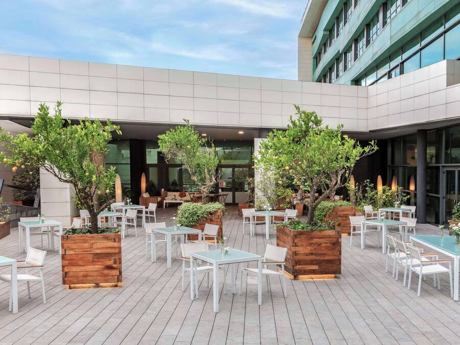 An outdoor patio area with white tables and chairs, surrounded by wooden planters with trees and plants.