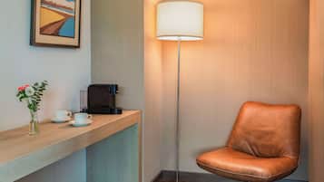A hotel reception area with a wooden counter, a coffee machine, a framed picture, a floor lamp, and a leather chair.