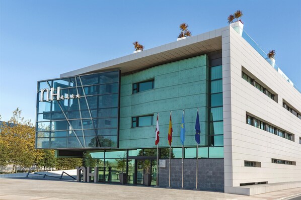 A modern building with a glass facade, multiple flags, and a clear sky. - NH Gijón (Gijon)