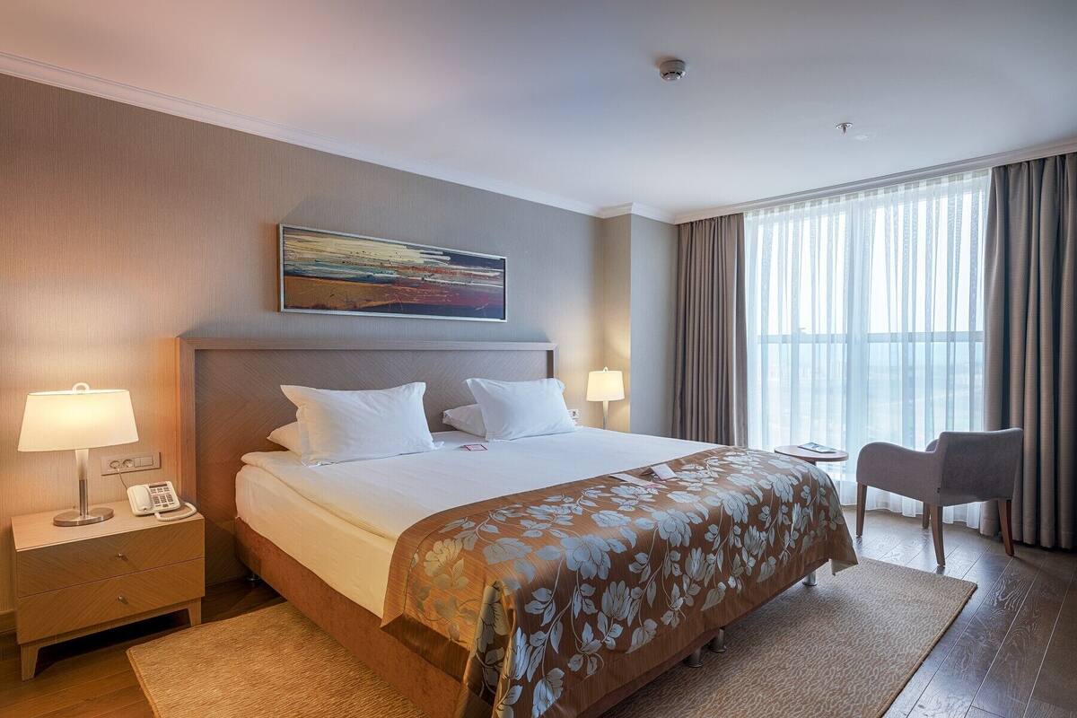 A hotel room with a large bed, two bedside tables with lamps, a chair, and a large window with curtains.