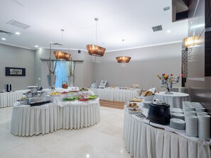 Un moderno salón de conferencias con mesas redondas cubiertas con manteles blancos, equipado con diversas estaciones de comida y bebidas.