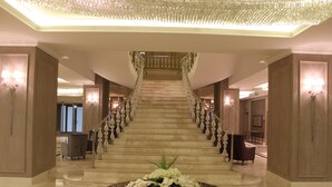 Una imponente escalera con revestimiento de mármol, flanqueada por elegantes apliques y un arreglo floral central sobre una mesa de mármol.