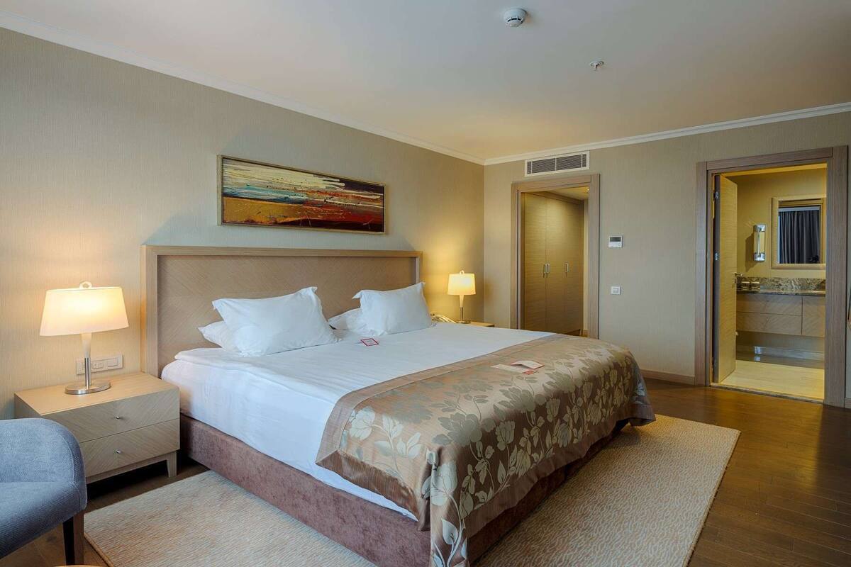 A hotel room with a large bed, two bedside lamps, a painting on the wall, and a door leading to a bathroom.
