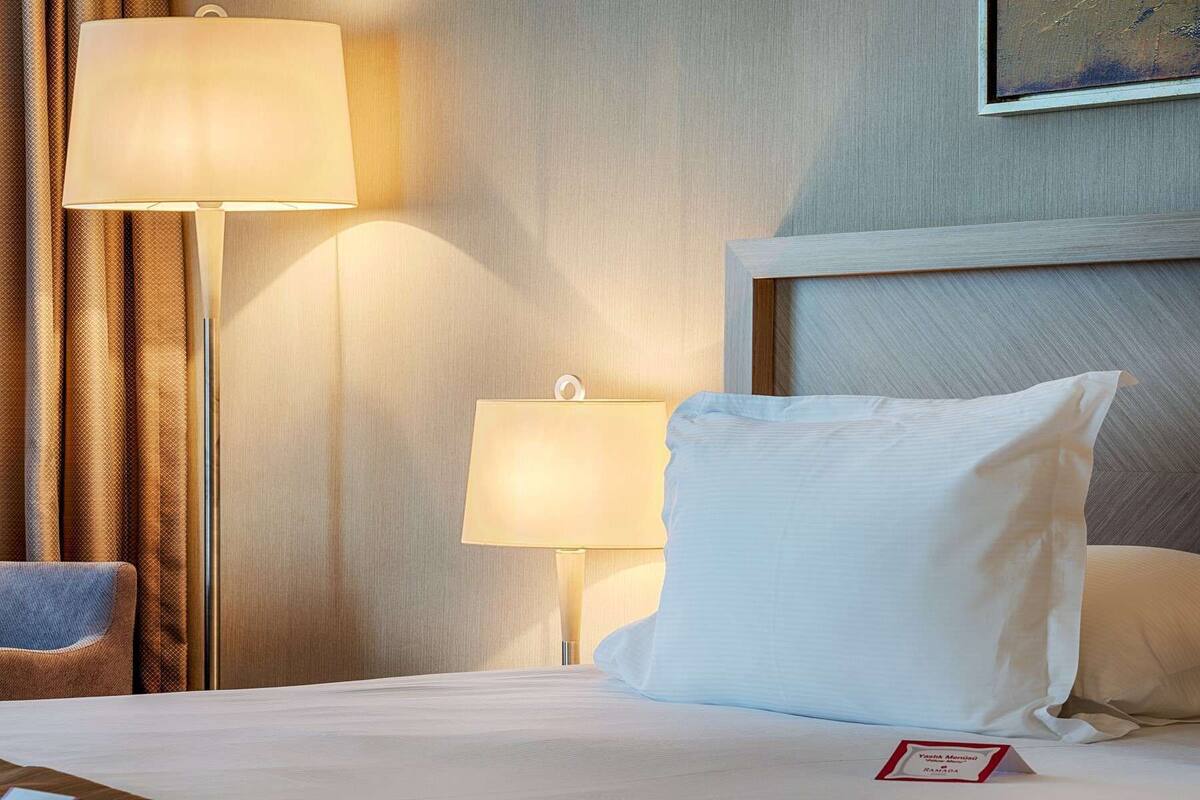 A hotel room with a bed, two bedside lamps, a headboard, and a framed picture on the wall.