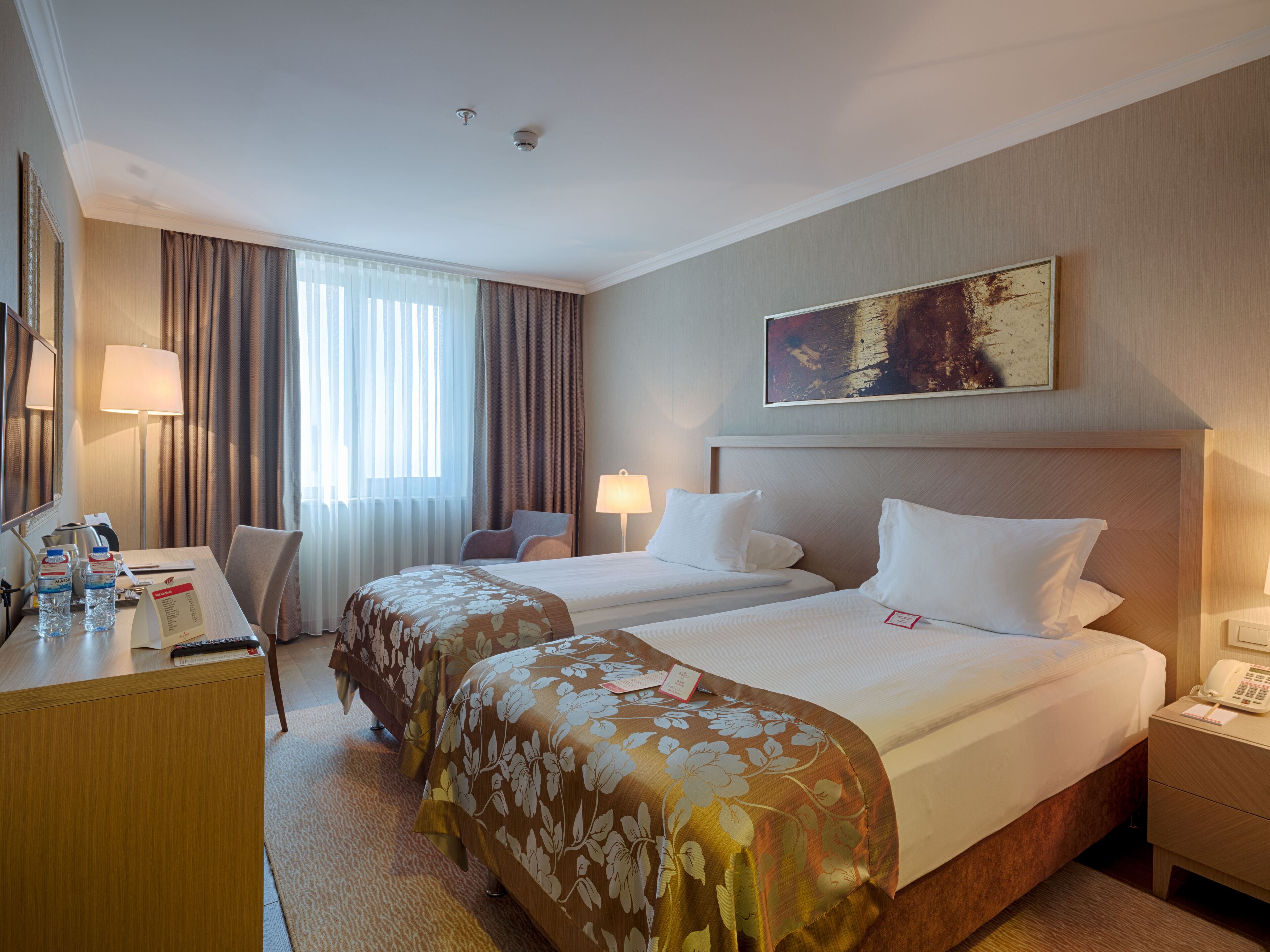 A hotel room with two beds, a desk with a lamp, a telephone, and a painting on the wall.