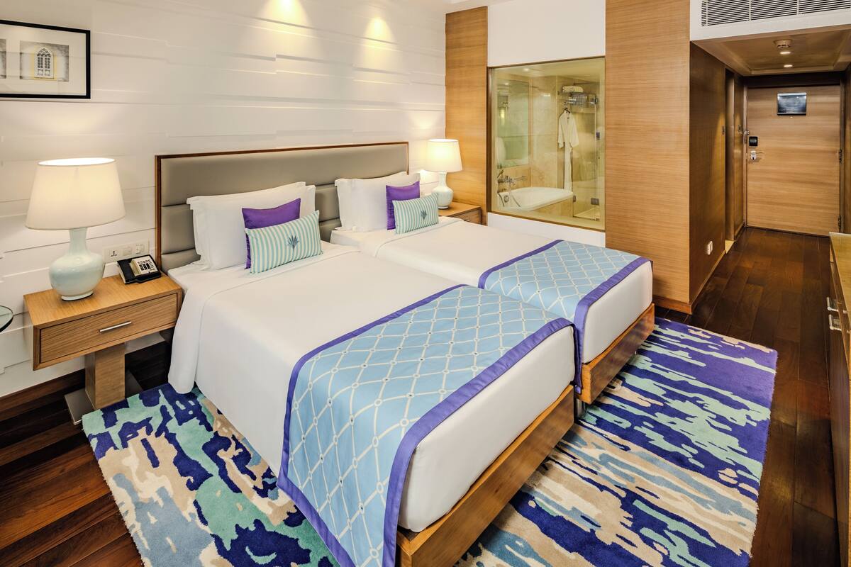 A hotel room with two beds, a bedside table with a lamp, a nightstand with a phone, a bathroom with a glass shower enclosure, and a wooden door.