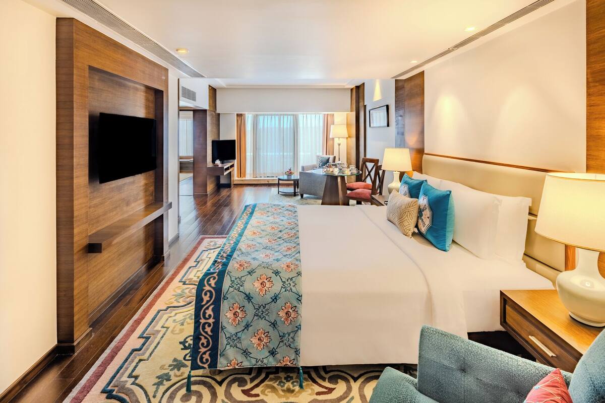 A hotel room with a large bed, a TV mounted on the wall, a desk with a chair, and a view of the city through the window.