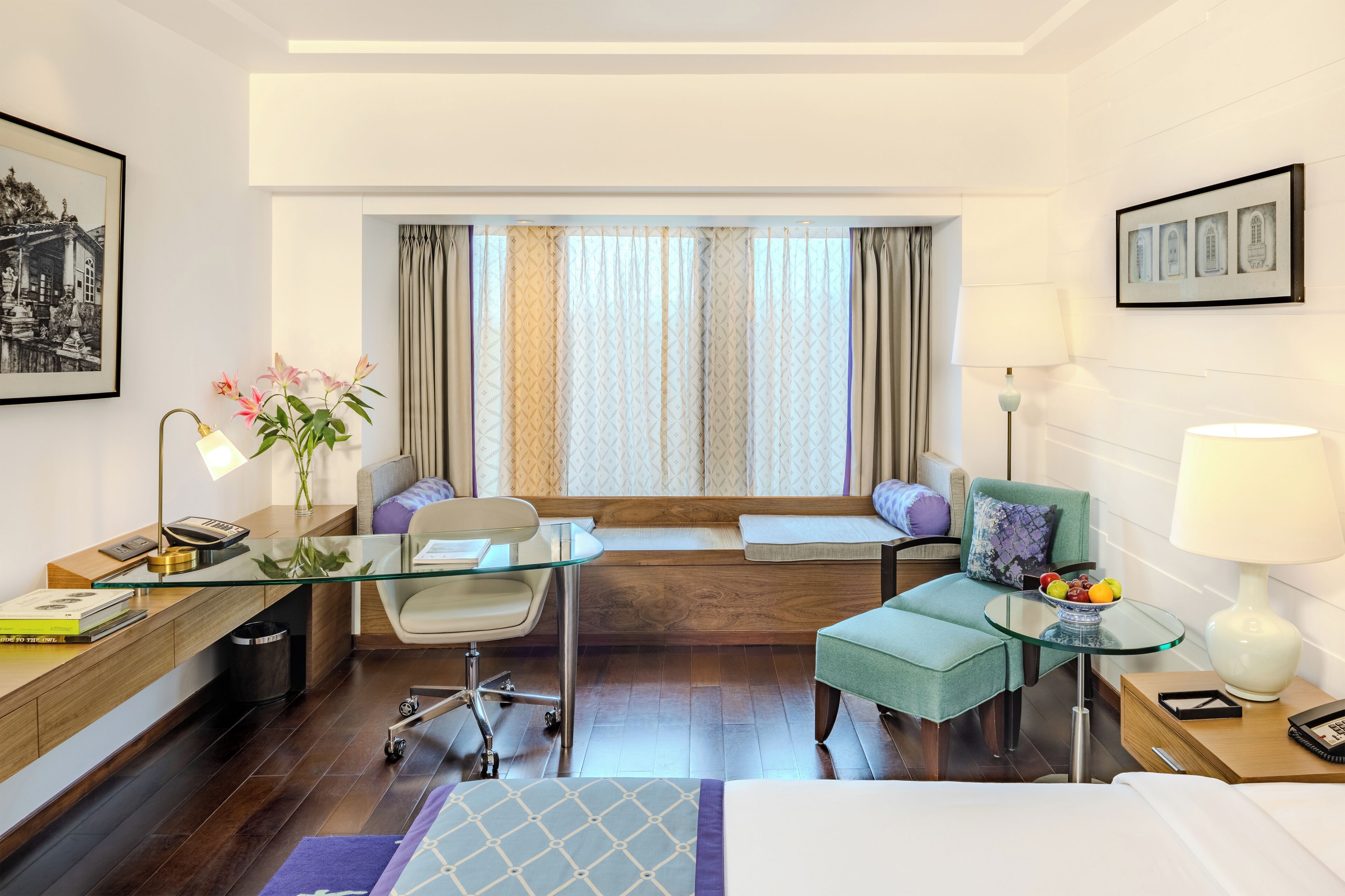 Habitación de hotel con un escritorio de vidrio, un sillón verde, un sofá, una cama y una ventana con cortinas.