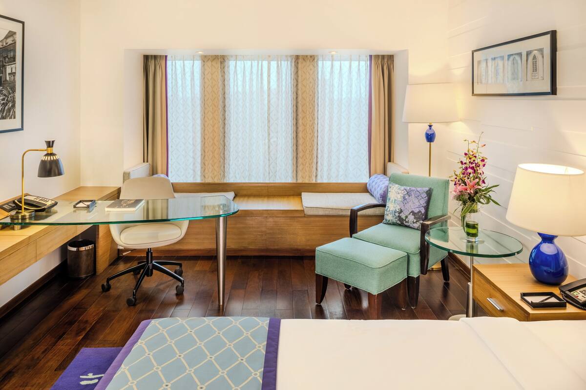 A hotel room with a glass desk, a green armchair, a bed, a window with curtains, and a vase with flowers.